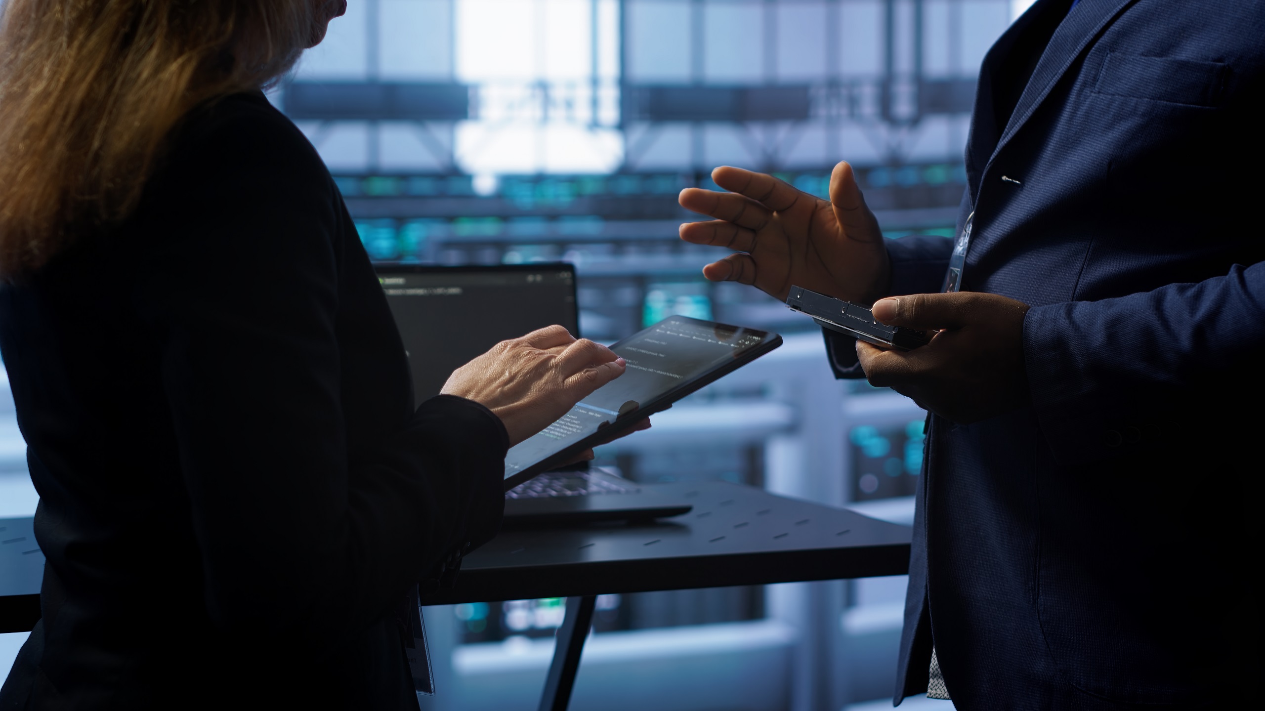 Two professionals in a data center discussing information on a tablet, with computer servers and monitors visible in the background.