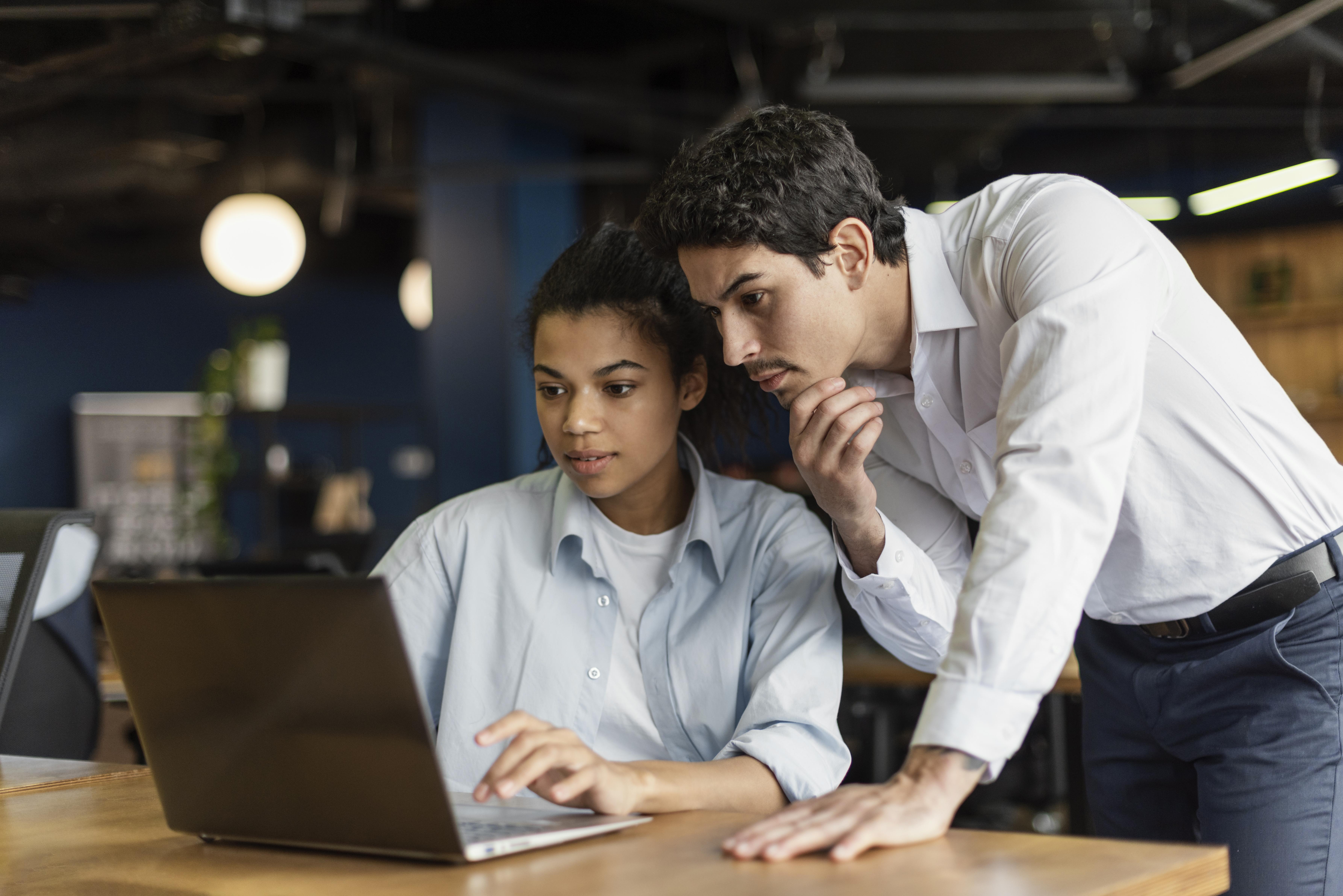 Two coworkers in an office collaborating at a desk, with one seated using a laptop and the other standing beside them looking at the screen.