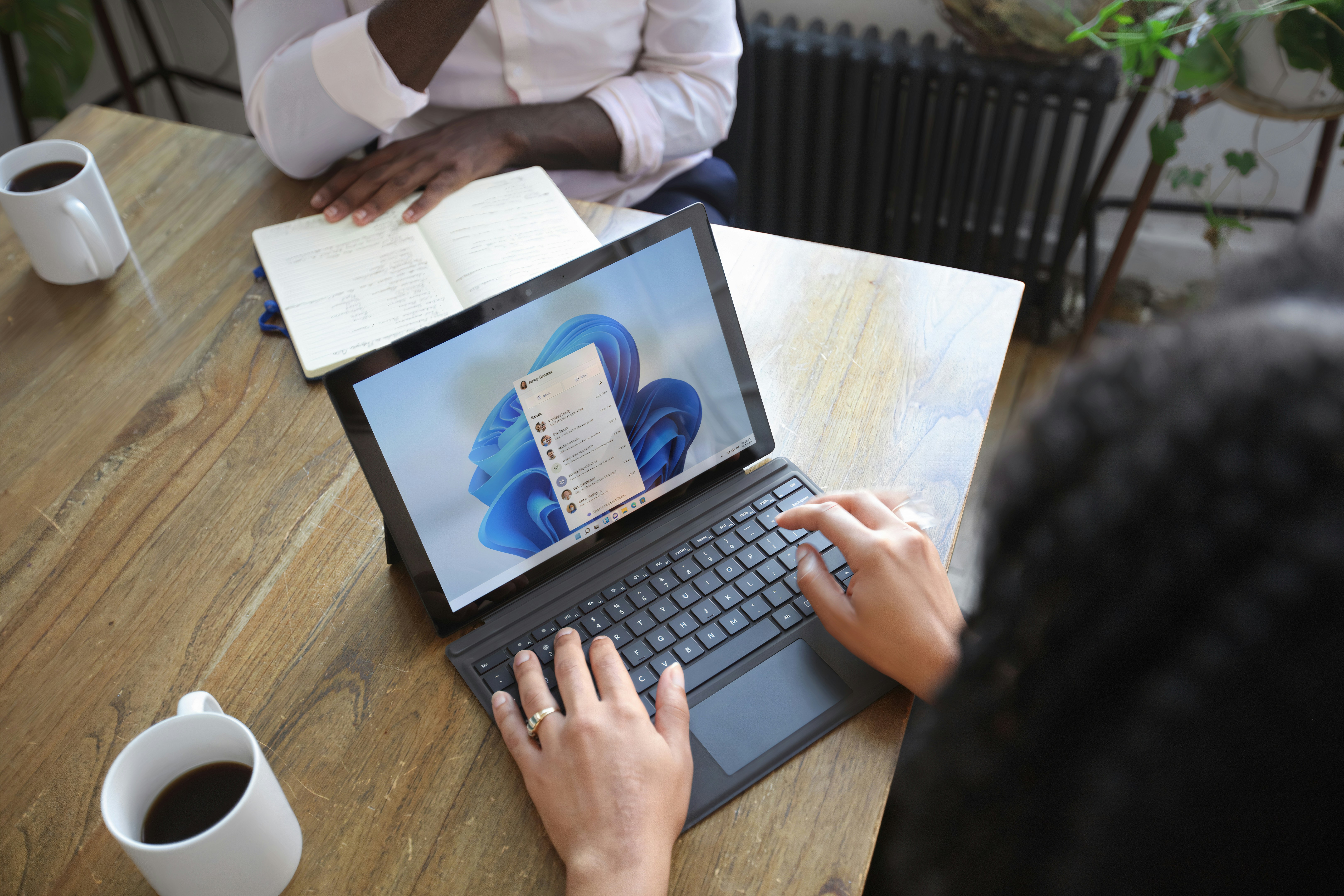 Tablet computer displaying Windows 11 desktop on a wooden table with a notebook and coffee cup