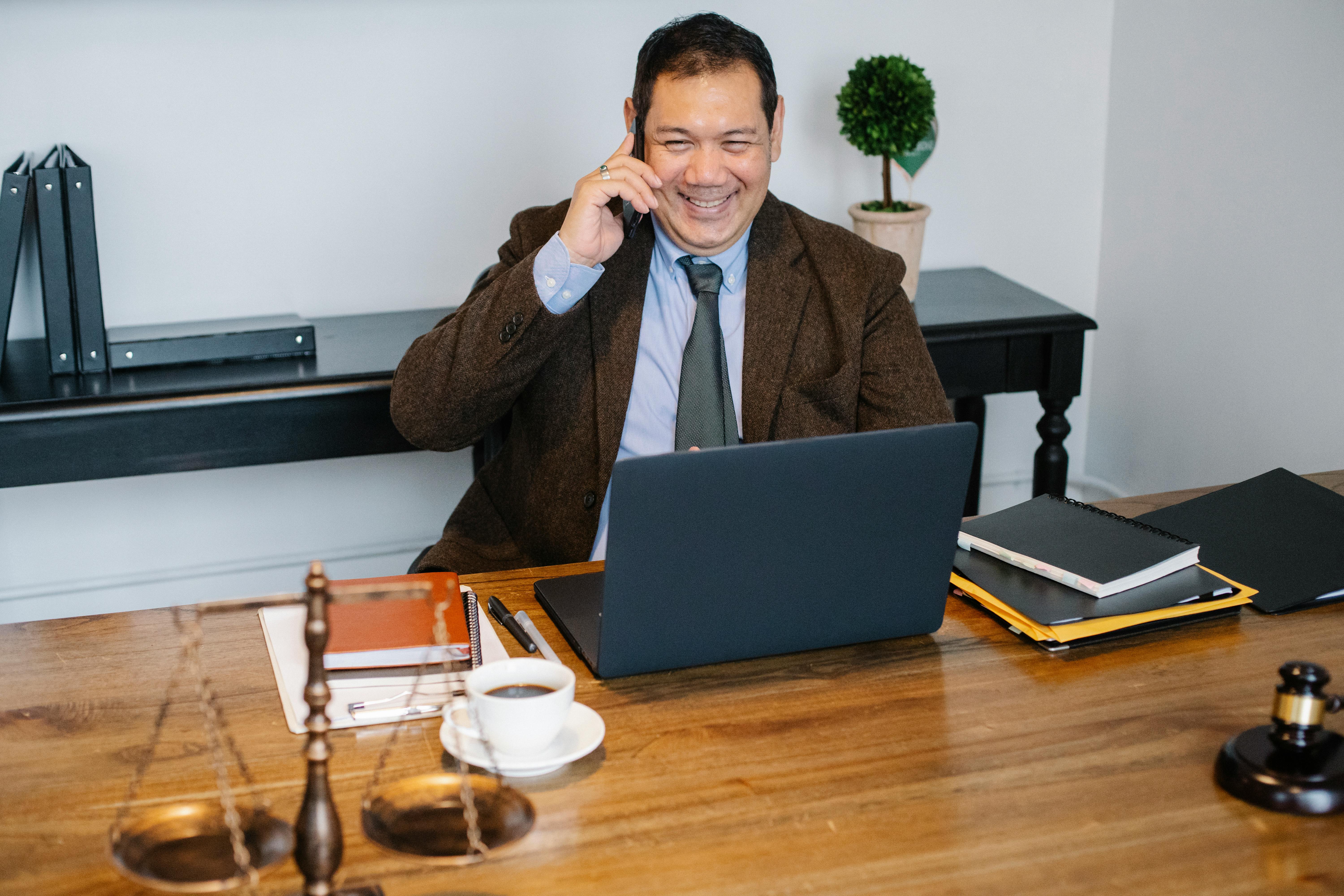Lawyer smiling while on a call in front of laptop, scale, notebooks, and coffee
