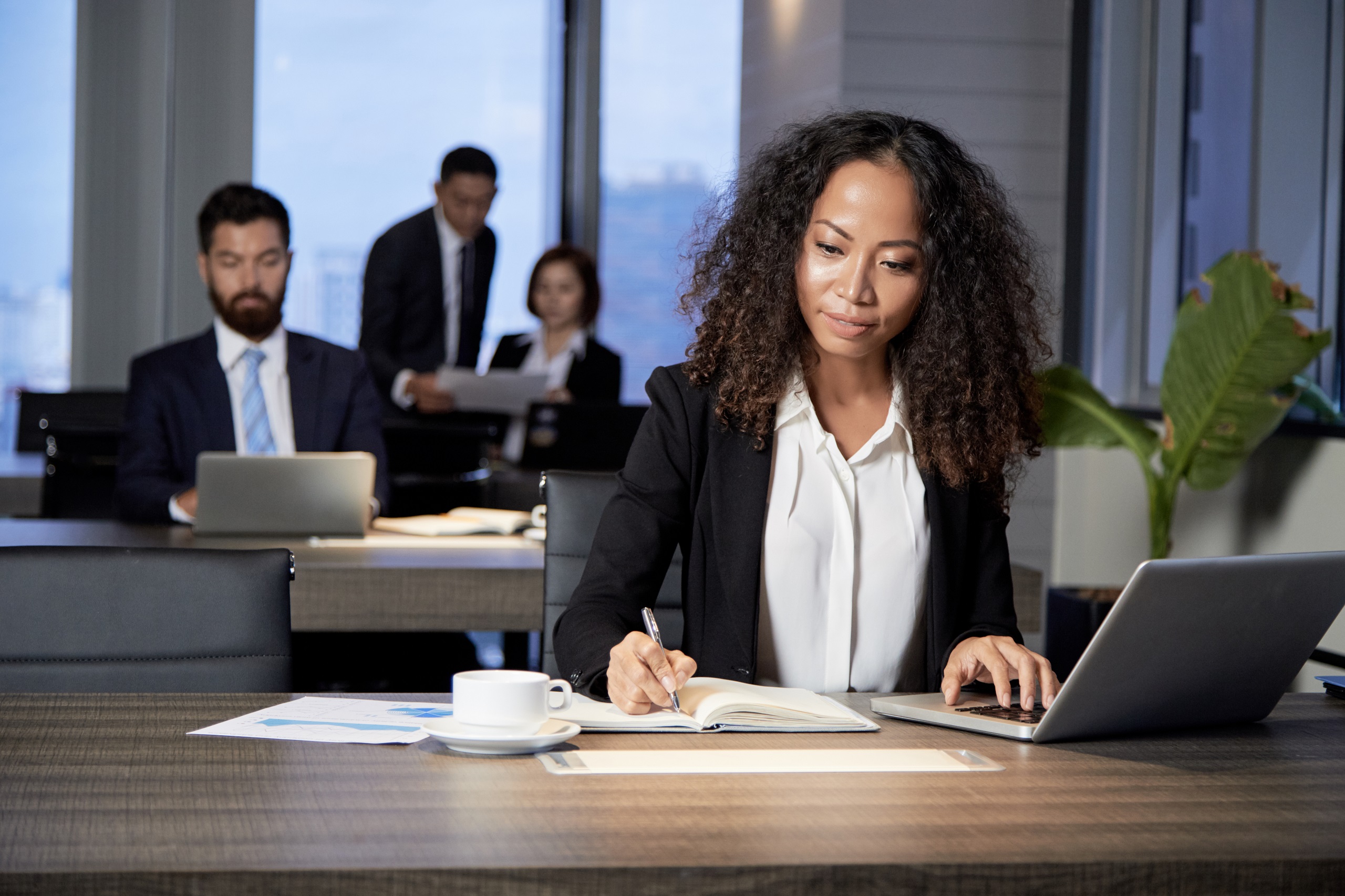 Woman in an office with coworkers typing on laptop and writing with pen in notebook