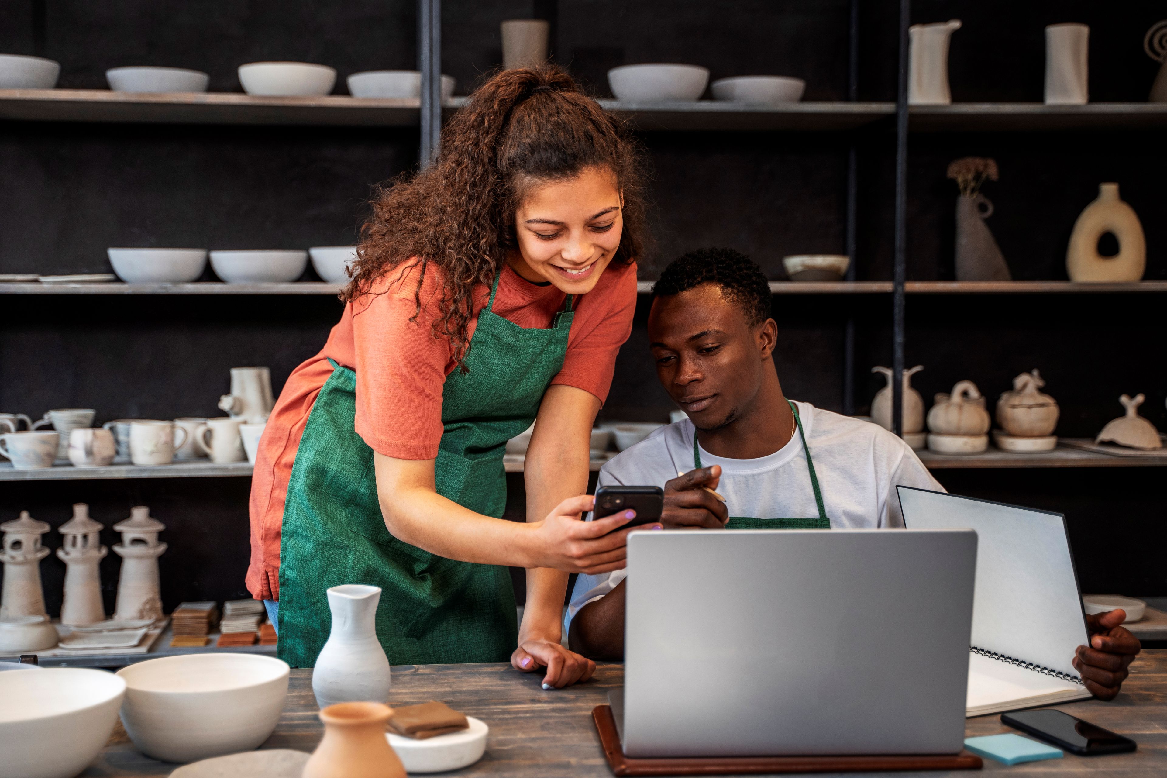 Two people looking at phone with laptop in a pottery studio.